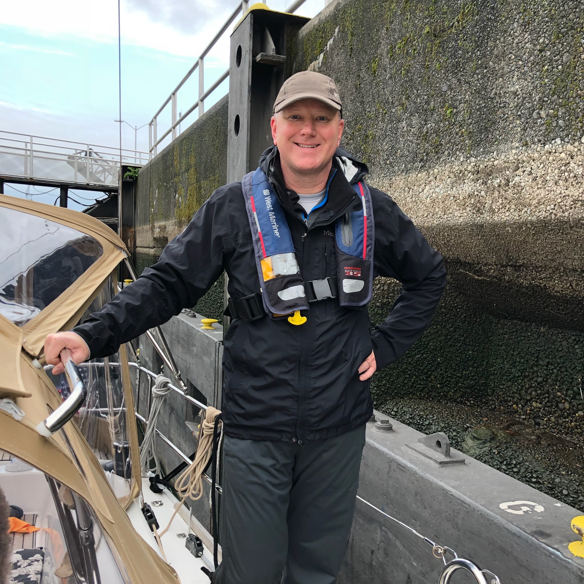 A sailor smiling at the helm beside a dock wall.