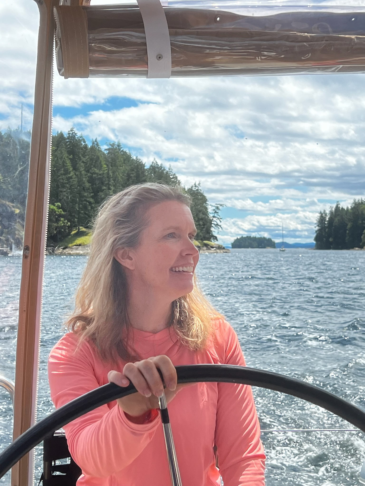A sailor in a coral shirt at the helm near wooded shoreline.